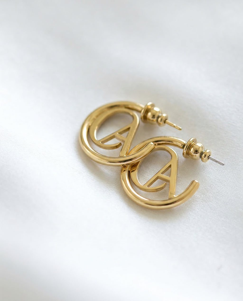 Gold ChristAdorn logo hoop earrings with polished finish and post-back closure on a white satin background