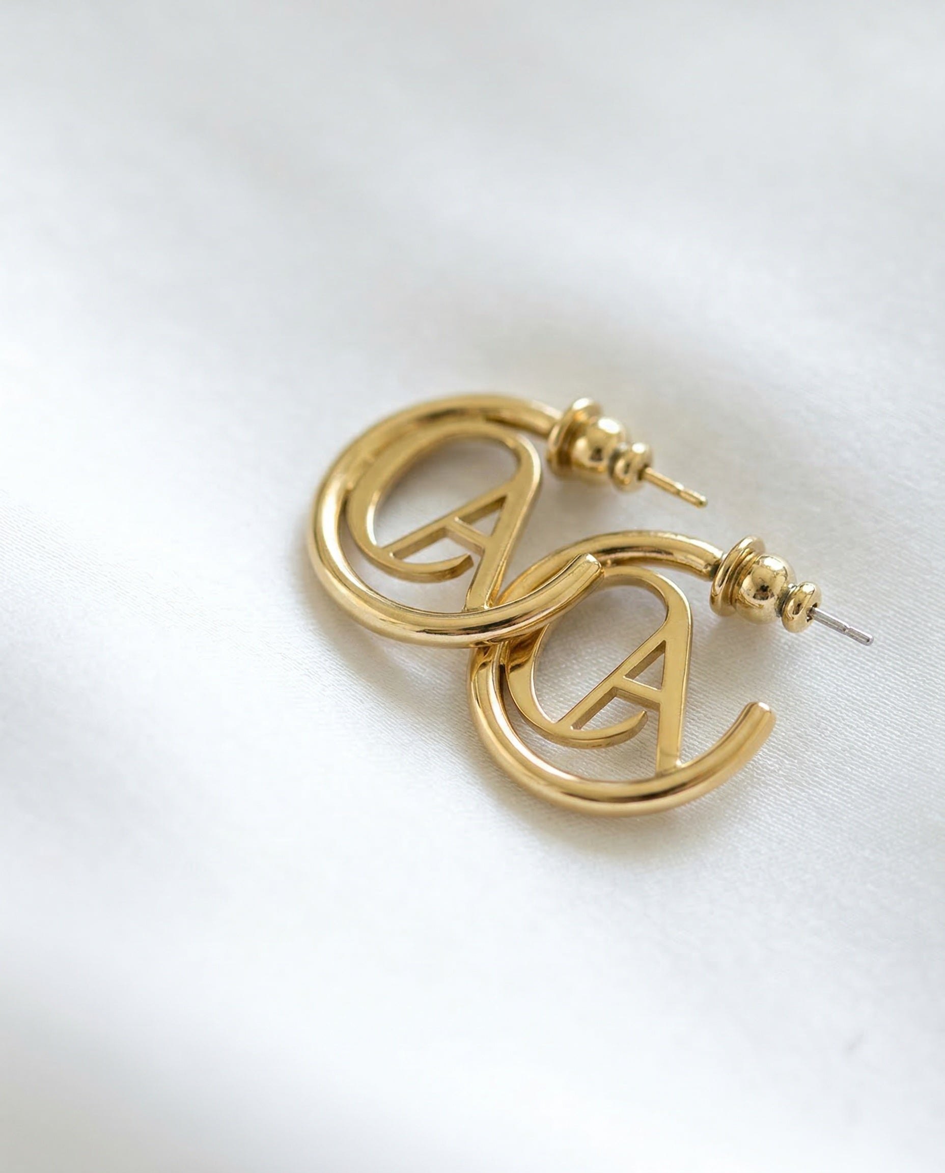 Gold ChristAdorn logo hoop earrings with polished finish and post-back closure on a white satin background