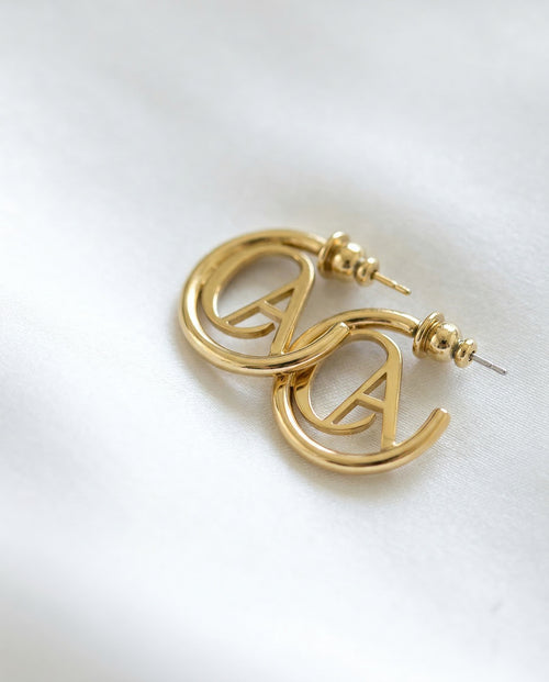 Gold ChristAdorn logo hoop earrings with polished finish and post-back closure on a white satin background